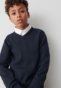 Tmavomodrý sveter s výstrihom do V z pletenej látky, s dlhými rukávmi a rebrovanými manžetami, nosený cez bielu košeľu s golierom.