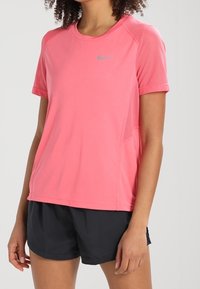 Rosa kortärmad träningsskjorta med nätaccenter, rund halsringning och en silverfärgad Nike-logga på övre vänster sida. Matchas med svarta shorts.