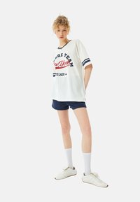 Camiseta blanca de gran tamaño con acentos en negro y azul marino, texto rojo que dice "Equipo Universitario Nueva York Est. 2020", combinada con pantalones cortos azul marino y zapatillas deportivas blancas.