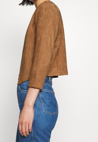 Veste en daim marron au design court, col rond et manches trois-quarts, associée à un jean denim bleu taille haute.