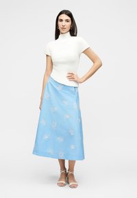 Γυναίκα στέκεται με το χέρι στη μέση, φορώντας λευκό πλεκτό κοντομάνικο τοπ, μπλε φλοράλ φούστα σε σχήμα Α και λευκά πέδιλα με λουράκια.