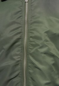 Fermeture éclair métallique argentée centrée sur un tissu matelassé vert olive, montrant des coutures et de légers plis sur les deux côtés.