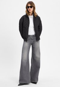 Giacca nera trapuntata con colletto alto, camicia bianca con bottoni e jeans grigi a gamba larga. Gli occhiali da sole completano l'outfit.