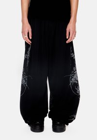 Άτομο που φορά χαλαρά μαύρα παντελόνια με λευκά αφηρημένα γραφικά σχεδιασμένα με γραμμές και μαύρα αθλητικά παπούτσια, στέκεται μπροστά σε λευκό φόντο.