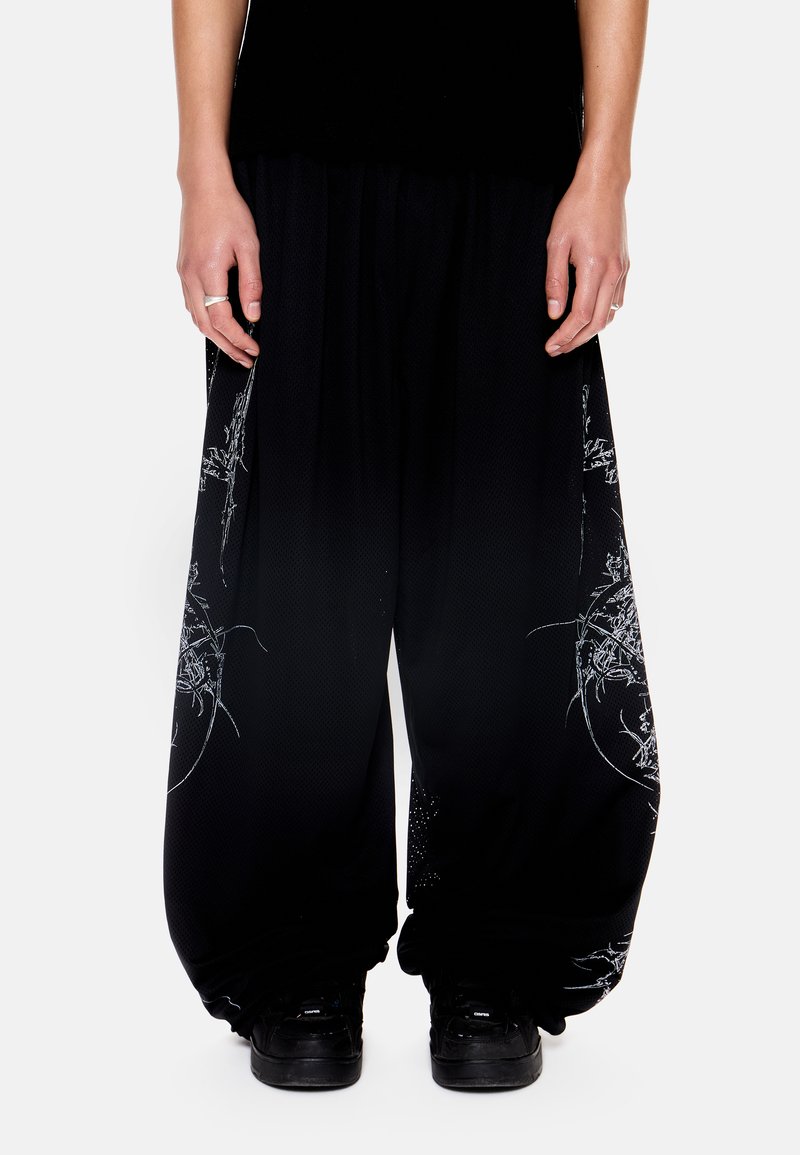 Άτομο που φορά χαλαρά μαύρα παντελόνια με λευκά αφηρημένα γραφικά σχεδιασμένα με γραμμές και μαύρα αθλητικά παπούτσια, στέκεται μπροστά σε λευκό φόντο.