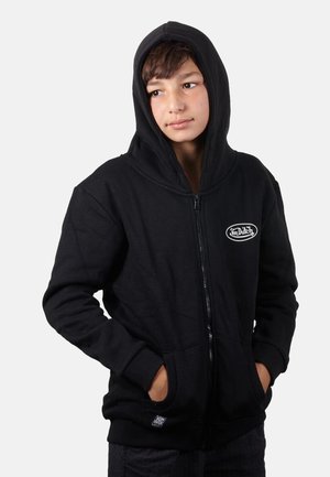 Garçon portant un sweat à capuche noir zippé, capuche relevée, mains dans les poches, regardant légèrement sur le côté sur un fond uni.