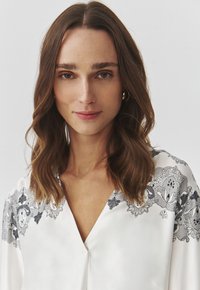 Joven mujer con cabello castaño ondulado a la altura de los hombros lleva una blusa blanca con patrones florales negros tipo encaje sobre un fondo claro.