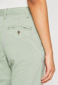 Prawa ręka osoby obok jasnooliwkowych spodni, pokazująca tylnią kieszeń z guzikiem i szlufkami na pasek.