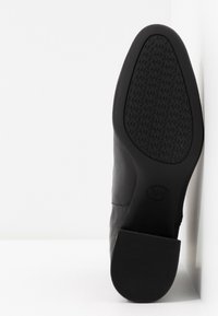 Semelle de botte en cuir noir avec une bande de roulement texturée et un logo imprimé, inclinée verticalement contre une surface blanche.
