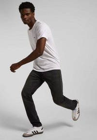 Camiseta blanca con logo negro, jeans oscuros y zapatillas grises con rayas negras; modelo en medio de un paso, mostrando un atuendo casual.