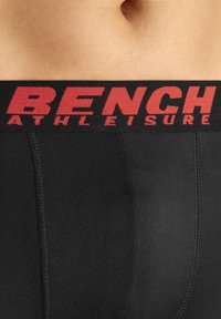 Black athletic shorts feature a wide elastic waistband with red "BENCH ATHLEISURE" text, smooth texture, and fitted design.