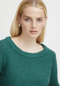 Grüner Strickpullover mit einem gerippten Ausschnitt und strukturiertem Stoff. Das Design verfügt über lange Ärmel und subtile Farbvariationen.