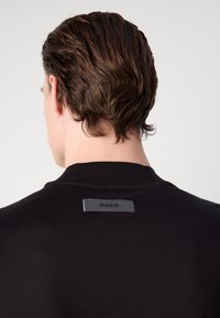 Černá bavlněná mikina s kulatým výstřihem a malou, obdélníkovou nálepkou "HUGO" na zadní straně, která kontrastuje s látkou.