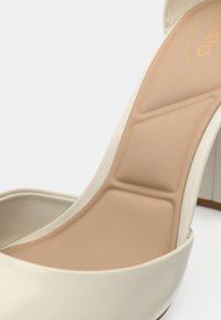 Korkokengät, joissa on beige pehmustettu sisäpohja ja kermaisen värinen ulkomateriaali, näyttäen yksityiskohtaisen ergonomisen pehmustuksen jalkatuen tueksi.