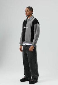 Grijs-zwart vest met rits en een witte afbeelding, gecombineerd met loszittende donkere jeans en zwarte plateauzolen met gekleurde veters.