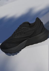 Sort chunky sneakers med tyk sål og sorte snørebånd, vist mod en blå og hvid baggrund med bløde skygger.