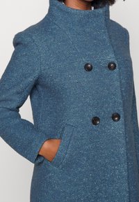 Kabát z vlněné směsi v tmavě modré barvě s texturovaným povrchem, s vysokým límcem, černými dvouřadovými knoflíky a bočními kapsami.