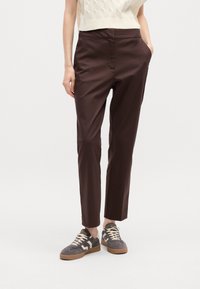 Brown tapered trousers made of smooth fabric with side pockets, paired with gray sneakers featuring striped accents and light laces.