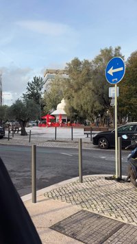 Rode parasols met witte tekst beschaduwen metalen stoelen en tafels op een plein. Achter bomen staat een wit prieel, met geparkeerde auto's langs de straat.