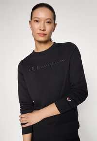 Črna majica s kapuco, izdelana iz mehkega materiala, z dvignjenim logotipom "Champion" v črni barvi, rebrastimi manšeti in klasičnim okroglim izrezom.