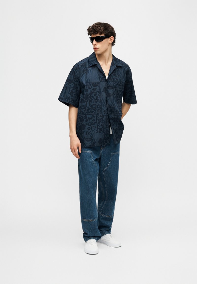 Giovane uomo con camicia nera a maniche corte con motivo, jeans blu larghi, sneakers bianche e occhiali da sole neri, in piedi con una mano nella tasca.