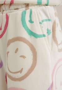 Vitt tyg med en mjuk textur som har färgglada mönster av glada ansikten i rosa, gröna, bruna och lila nyanser. Elastisk midjebanddetalj synlig.