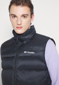 Černý bezrukávový péřový vestik s vysokým límcem, lesklou texturou, zipovým zapínáním a logem Columbia na levé hrudi.