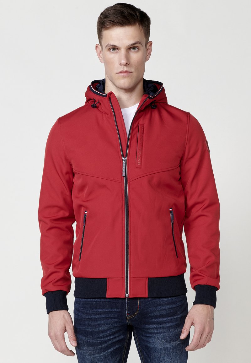 Rød lynlåsjacket med hætte, med en marineblå inderside, sidelommer og ribbede manchetter og kant for en tætsiddende pasform. Glat, mat stoftekstur.