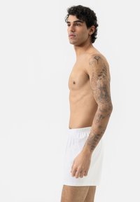 Shorts en coton blancs avec une taille élastique, présentés en profil latéral sur une personne avec des tatouages sur le bras. Texture lisse, design décontracté.