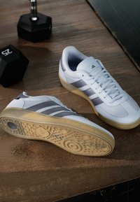 Witte sportschoenen met grijze strepen, met een gestructureerde rubberen zool en veters, geplaatst op een houten oppervlak naast een dumbbell.