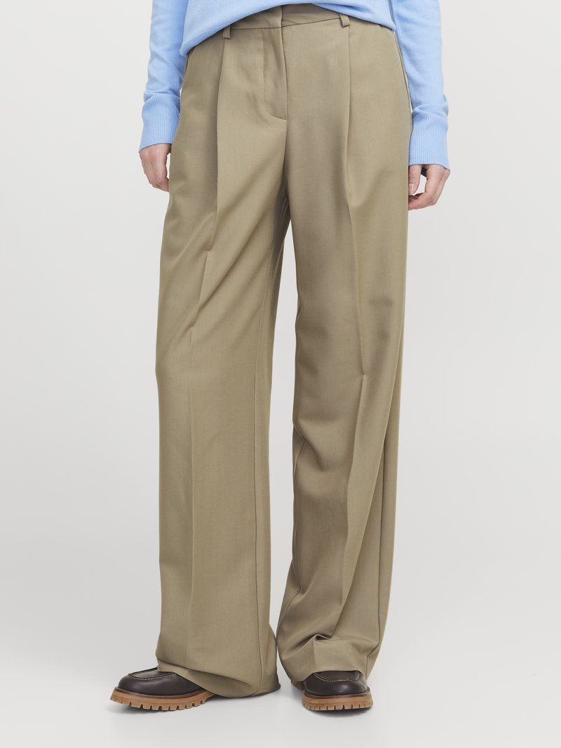 Beige weit geschnittene Hose aus einem glatten Stoff, mit Falten und hoher Taille. Kombiniert mit dunkelbraunen Schnürschuhen.