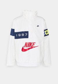 Chaqueta exterior blanca con cuello alto, logo rojo de Nike, detalles en azul, gráfico "1987", cordón ajustable y puños elásticos.