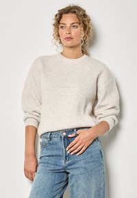 Sweter w kolorze kremowym, o dzianinowym splocie z prążkowanym wzorem, okrągłym dekoltem i opuszczonym rękawami, zestawiony z jasnoniebieskimi dżinsami.
