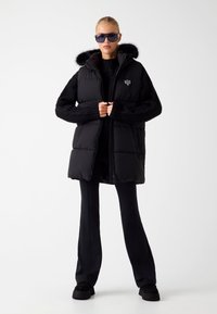 Juodas pūkinis liemenė su kapišonu, pūkų apvadu ir siuvinėtu raštu. Dėvima virš juodos megztos švarkelės, derinama su juodomis plačiomis kelnėmis ir bateliais.