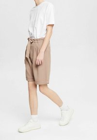 Pantalones cortos beiges de cintura alta, ajustados, con un lazo frontal y puños doblados, combinados con una camiseta blanca lisa y zapatillas blancas.