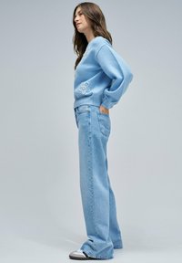 Suéter azul claro con bordado floral, combinado con jeans azul claro de corte holgado y zapatillas blancas, sobre un fondo neutral.