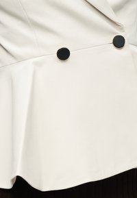 Veste blanc cassé à coupe péplum, dotée de deux boutons ronds noirs et d'une texture de tissu lisse. Détail de couture visible.