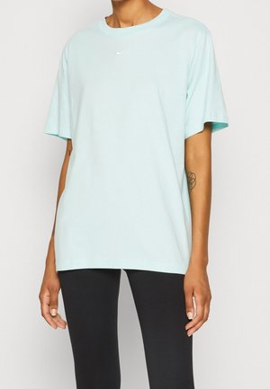 Persoon draagt een loszittend lichtblauw Nike-T-shirt en zwarte broek, met een tatoeage zichtbaar op de rechter onderarm.