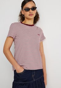 Νεαρή γυναίκα που φοράει μαύρα γυαλιά ηλίου, χρυσούς κρίκους στα αυτιά, ριγέ μαρουνί και λευκή μπλούζα, και σκουρόχρωμη τζιν φούστα με το χέρι στην τσέπη.