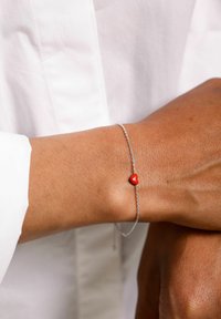 Silversmyckesarmband med en liten röd hjärtcharm, bärs på en handled mot en vit bakgrund. Armbandet har en delikat design.