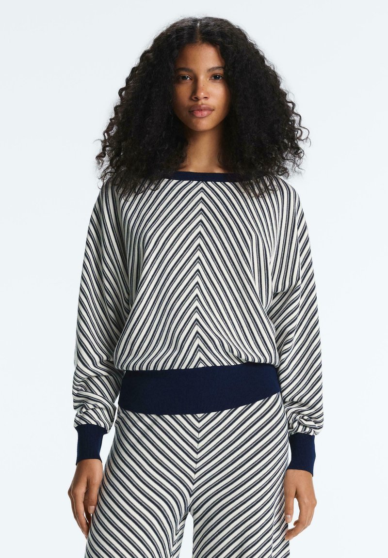 Femme aux cheveux bouclés portant un ensemble assorti de pull et pantalon rayés bleu marine et blanc, debout devant un fond clair uni.
