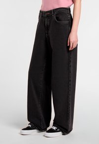 Black, wide-leg jeans made of denim, featuring a high waist, front pockets, and a subtle texture. Styled with black sneakers.