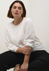 White, soft knit sweater with a round neckline and three-quarter sleeves. Worn with black pants, featuring ribbed cuffs and hem.