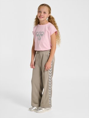 Jong meisje met krullend haar dat een roze t-shirt met een bij-logo draagt, beige trainingsbroek met chevronstrepen en zilveren sneakers, staand ontspannen.