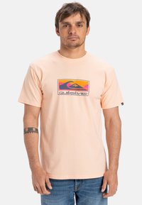 Tenký broskvový bavlněný tričko s barevným grafickým logem Quiksilver na hrudi. Krátké rukávy a volný střih.