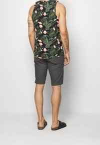 Top negro sin mangas con un patrón de hoja tropical verde y flores rosadas, combinado con pantalones cortos grises. Modelo de espaldas, llevando sandalias marrones.