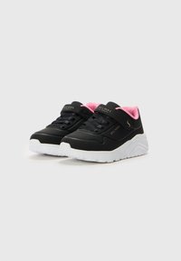 Baskets noires avec une texture lisse, dotées d'une doublure intérieure rose, d'une semelle blanche et d'une sangle en velcro sur les lacets pour un ajustement sécurisé.