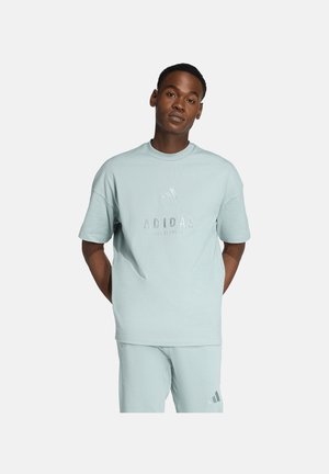 Ein Mann steht mit den Händen hinter dem Rücken, trägt ein hellblaues Adidas-T-Shirt und passende Shorts vor einem einfarbigen Hintergrund.