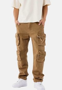 Pantalones cargo marrones hechos de una tela resistente, con múltiples bolsillos y cremalleras. Tienen un corte holgado y se llevan con zapatillas blancas.