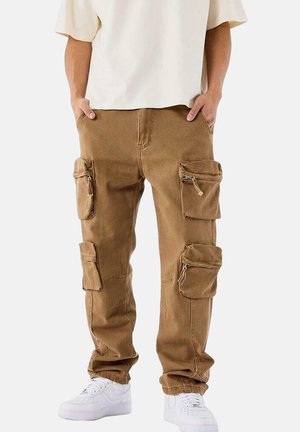 Pantalones cargo marrones hechos de una tela resistente, con múltiples bolsillos y cremalleras. Tienen un corte holgado y se llevan con zapatillas blancas.